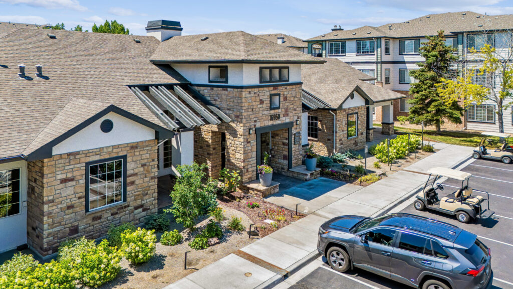 A stone-clad building with tall windows sits next to a parking lot with a black SUV and two golf carts. There are bushes, small trees, and sidewalks in front. Two-story buildings are in the background.