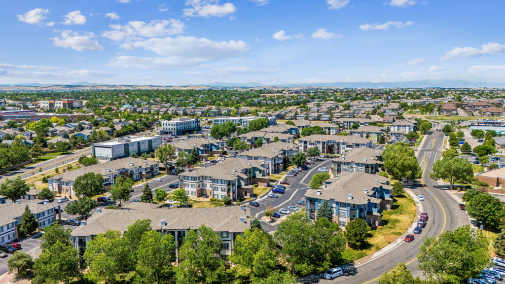 Aerial view of a suburban neighborhood with rows of apartment buildings, tree-lined streets, parked cars, and clear blue sky with scattered clouds in the background.