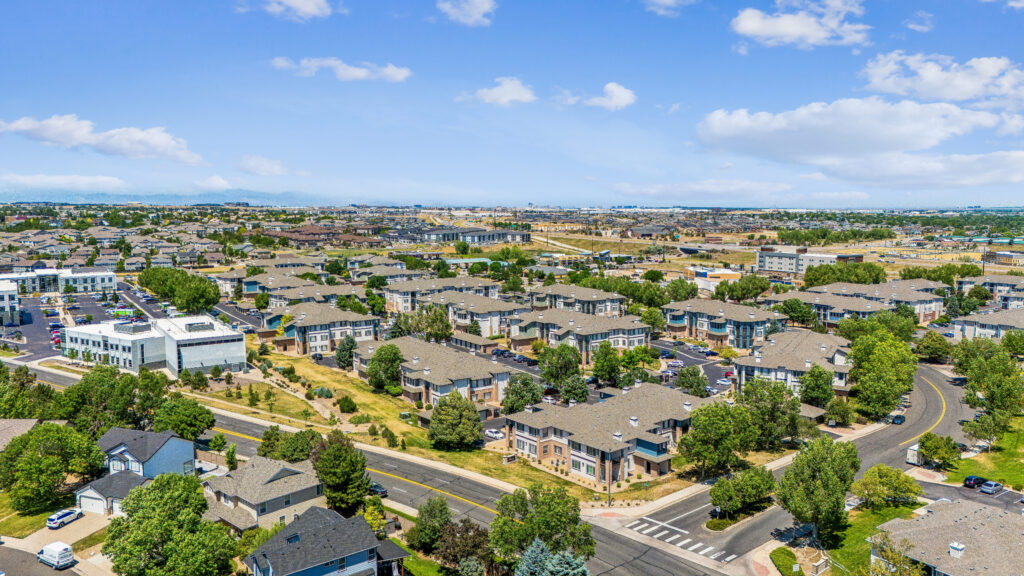 Aerial view of a suburban neighborhood with rows of houses, tree-lined streets, and some green spaces under a blue sky with scattered clouds.