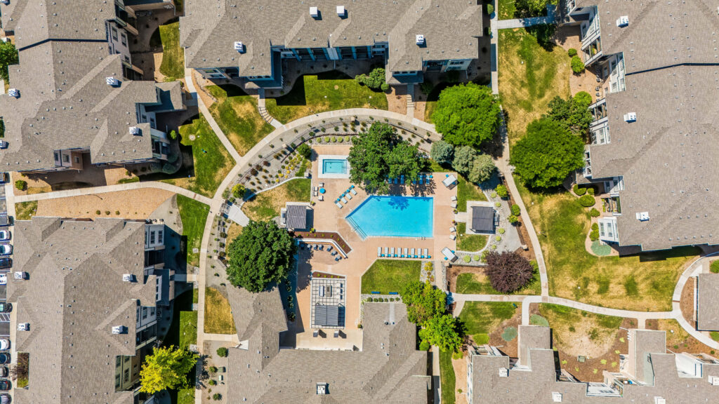 Aerial view of an apartment complex featuring a central swimming pool, lounge chairs, greenery, paths, and surrounding residential buildings.