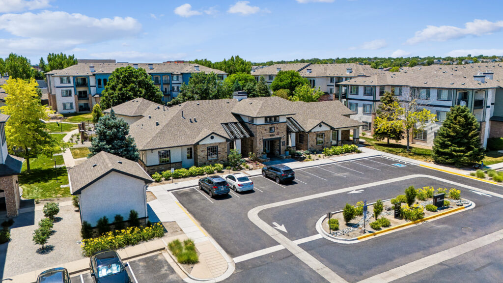 Aerial view of an apartment complex with multiple buildings, parked cars, landscaped greenery, and a clear blue sky in the background.