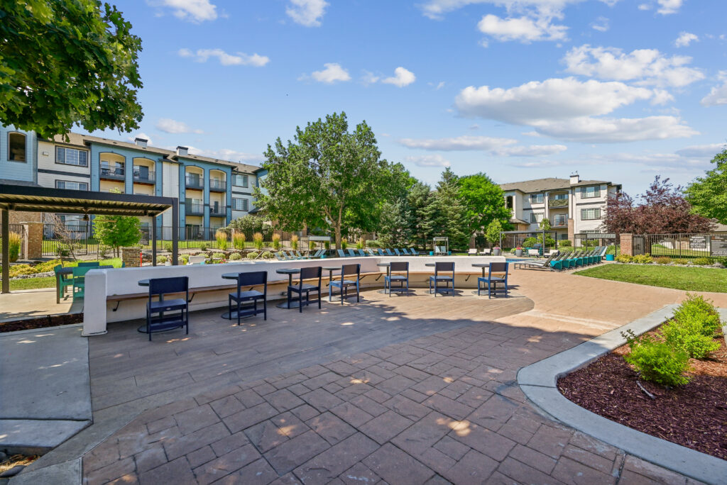 An outdoor patio area with a stone-topped bar, several blue chairs, and a paved walkway. In the background, there is a swimming pool with lounge chairs surrounded by multi-story apartment buildings, green trees, and a blue, partly cloudy sky.