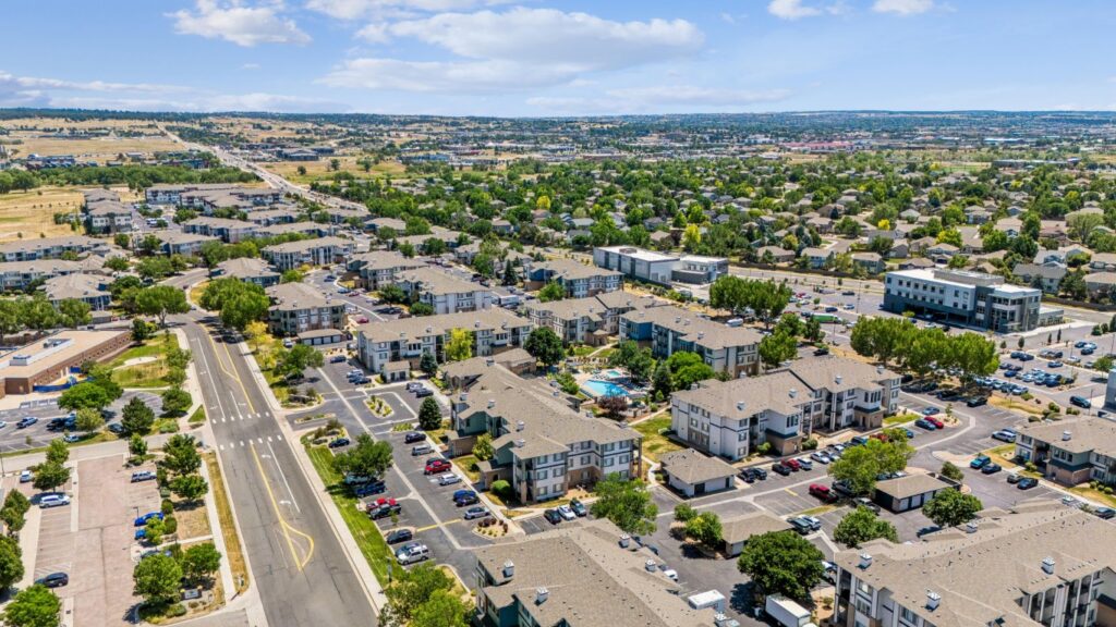Aerial view of a suburban neighborhood with apartment buildings, parking lots, trees, and roads, surrounded by greenery and distant hills under a partly cloudy sky.