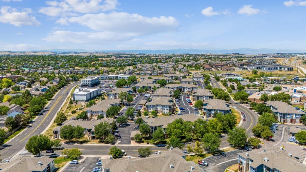 Aerial view of a suburban neighborhood with rows of houses, streets, trees, parking lots, and distant hills under a blue sky with scattered clouds.