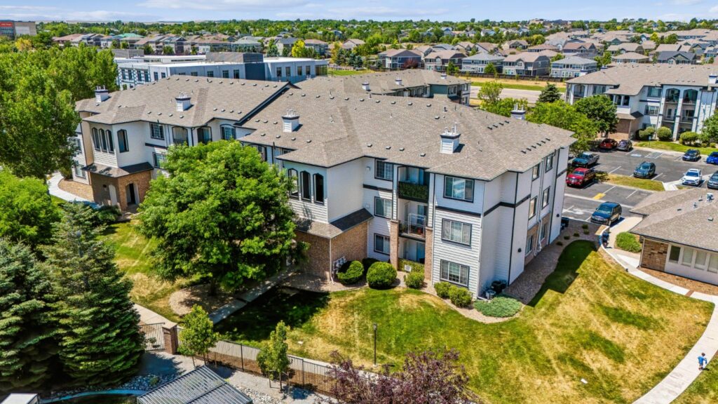 Aerial view of a multi-story apartment building with beige walls and gray roof, surrounded by green lawns, trees, and a parking lot with several parked cars. Other residential buildings are visible in the background.