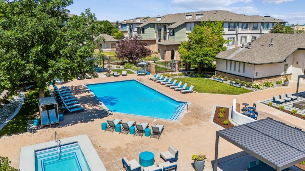 Aerial view of an outdoor swimming pool area with lounge chairs, hot tub, shaded seating, and landscaped greenery surrounded by apartment buildings.