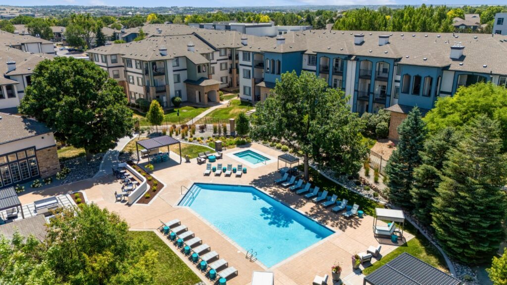 Aerial view of an apartment complex courtyard featuring a large, rectangular outdoor pool, lounge chairs, umbrellas, greenery, and nearby multi-story residential buildings under a clear sky.