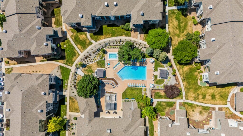 Aerial view of an apartment complex with a central pool, lounge chairs, a smaller hot tub, shaded seating areas, green trees, and walking paths surrounded by several buildings.